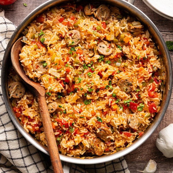 Overhead view of sausage, peppers, and rice skillet garnished with fresh parsley, shown in a stainless steel pan with a wooden spoon.