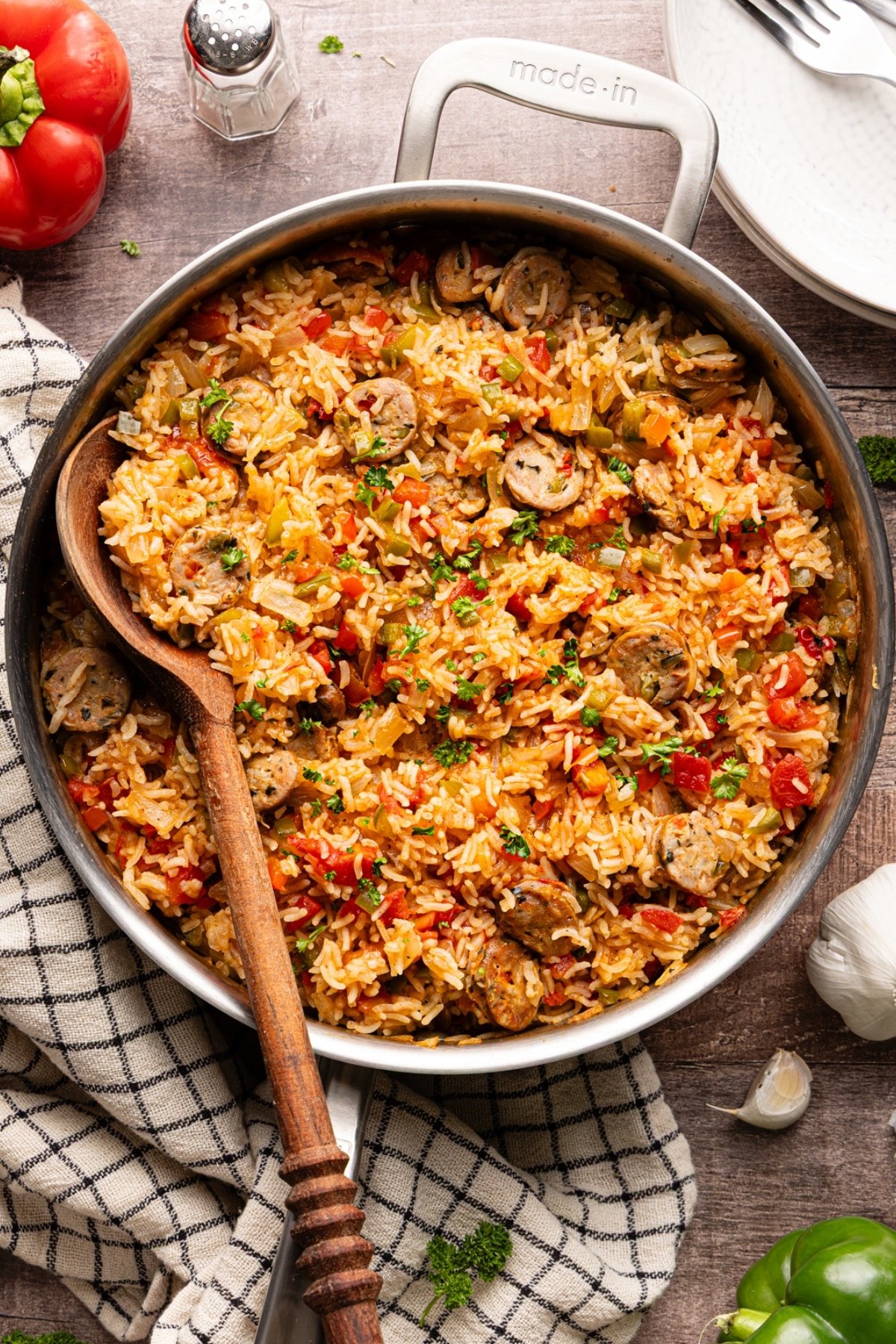 Overhead view of sausage, peppers, and rice skillet garnished with fresh parsley, shown in a stainless steel pan with a wooden spoon.