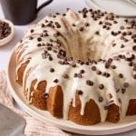 Chocolate chip Bundt cake topped with vanilla bean glaze and mini chocolate chips, displayed on a serving platter with a coffee mug and cake server nearby.