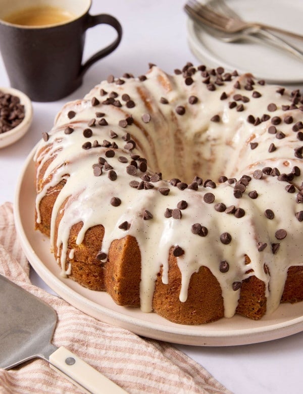 Chocolate chip Bundt cake topped with vanilla bean glaze and mini chocolate chips, displayed on a serving platter with a coffee mug and cake server nearby.