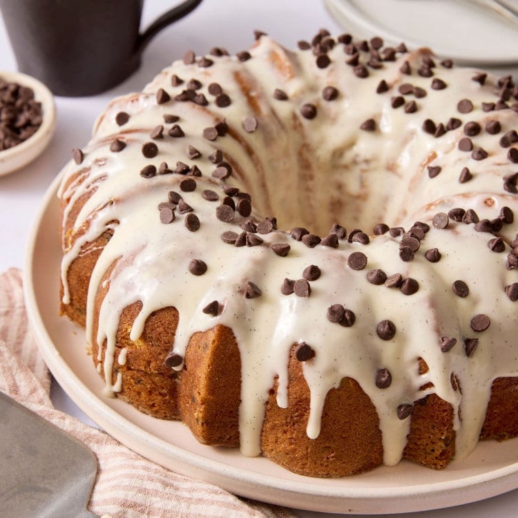 Chocolate chip Bundt cake topped with vanilla bean glaze and mini chocolate chips, displayed on a serving platter with a coffee mug and cake server nearby.