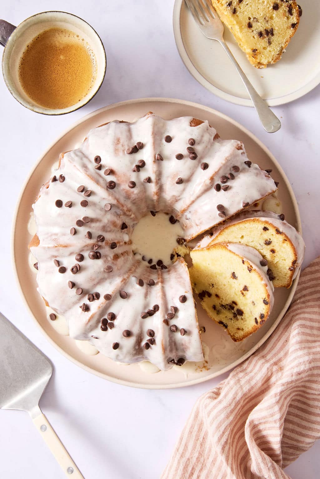 Overhead view of a chocolate chip Bundt cake covered with vanilla bean glaze and mini chocolate chips, displayed on a serving platter with sliced pieces ready to serve.