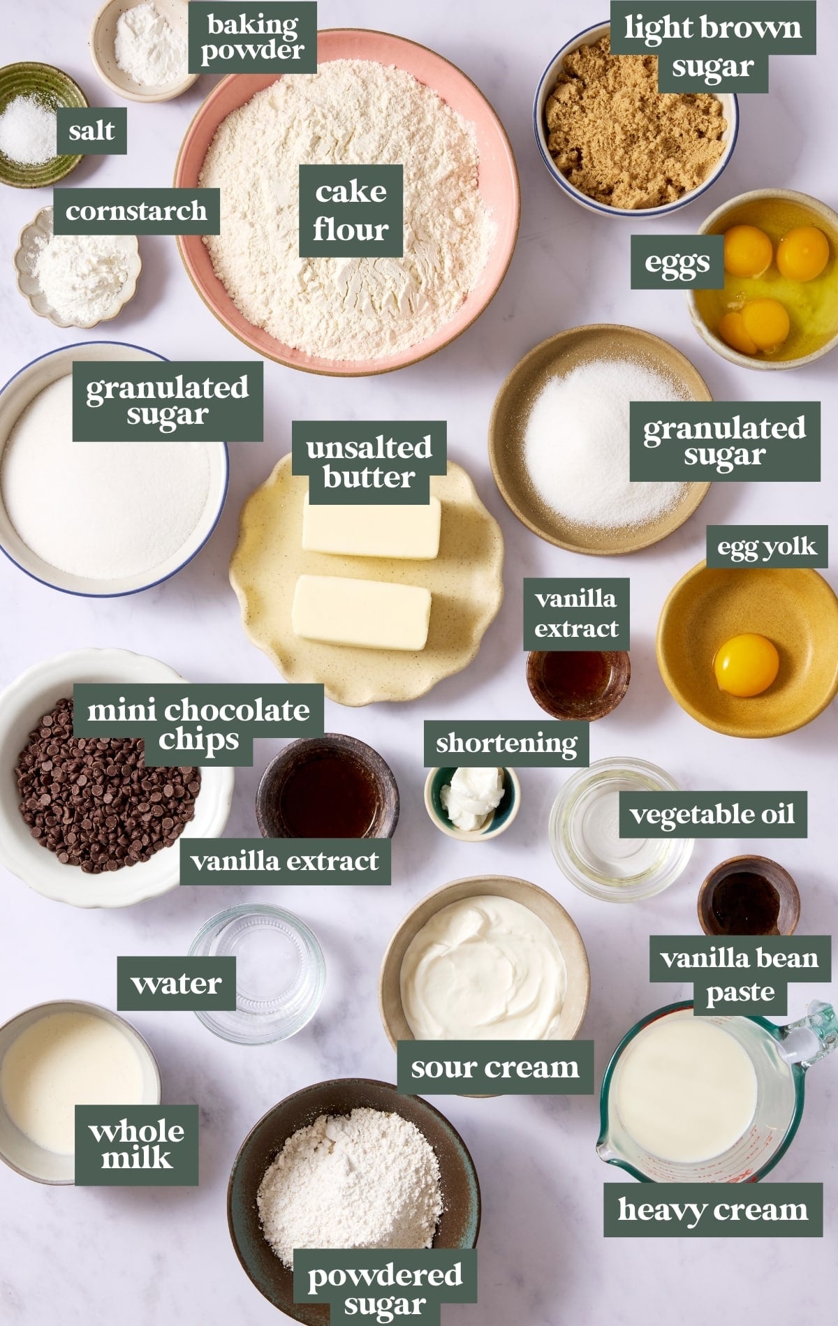 Overhead view of all ingredients needed for a chocolate chip Bundt cake arranged on a marble surface, including cake flour, cornstarch, baking powder, salt, granulated sugar, light brown sugar, butter, eggs, egg yolk, vanilla extract, vegetable oil, sour cream, whole milk, mini chocolate chips, powdered sugar, heavy cream, shortening, water, and vanilla bean paste, each labeled.