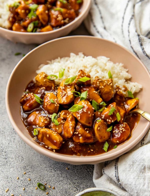 brown bowl with white rice, orange chicken, topped with chopped green onions and sesame seeds