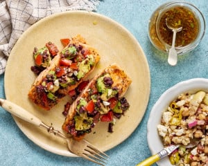 Mediterranean salmon topped with avocado tomato olive salsa served on a plate next to couscous salad and a jar of vinaigrette.