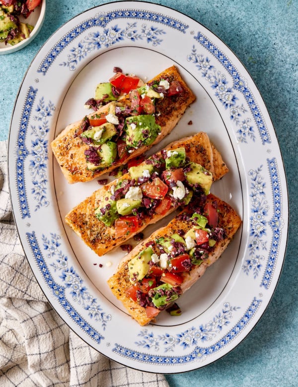 Mediterranean salmon fillets topped with avocado, tomato, olives, and feta arranged on a blue-and-white serving platter.