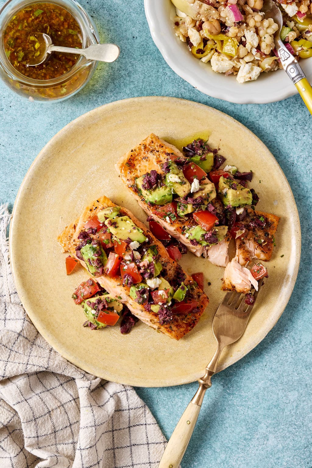 Two salmon fillets topped with avocado tomato olive salsa on a plate, with Mediterranean couscous salad and vinaigrette in the background.