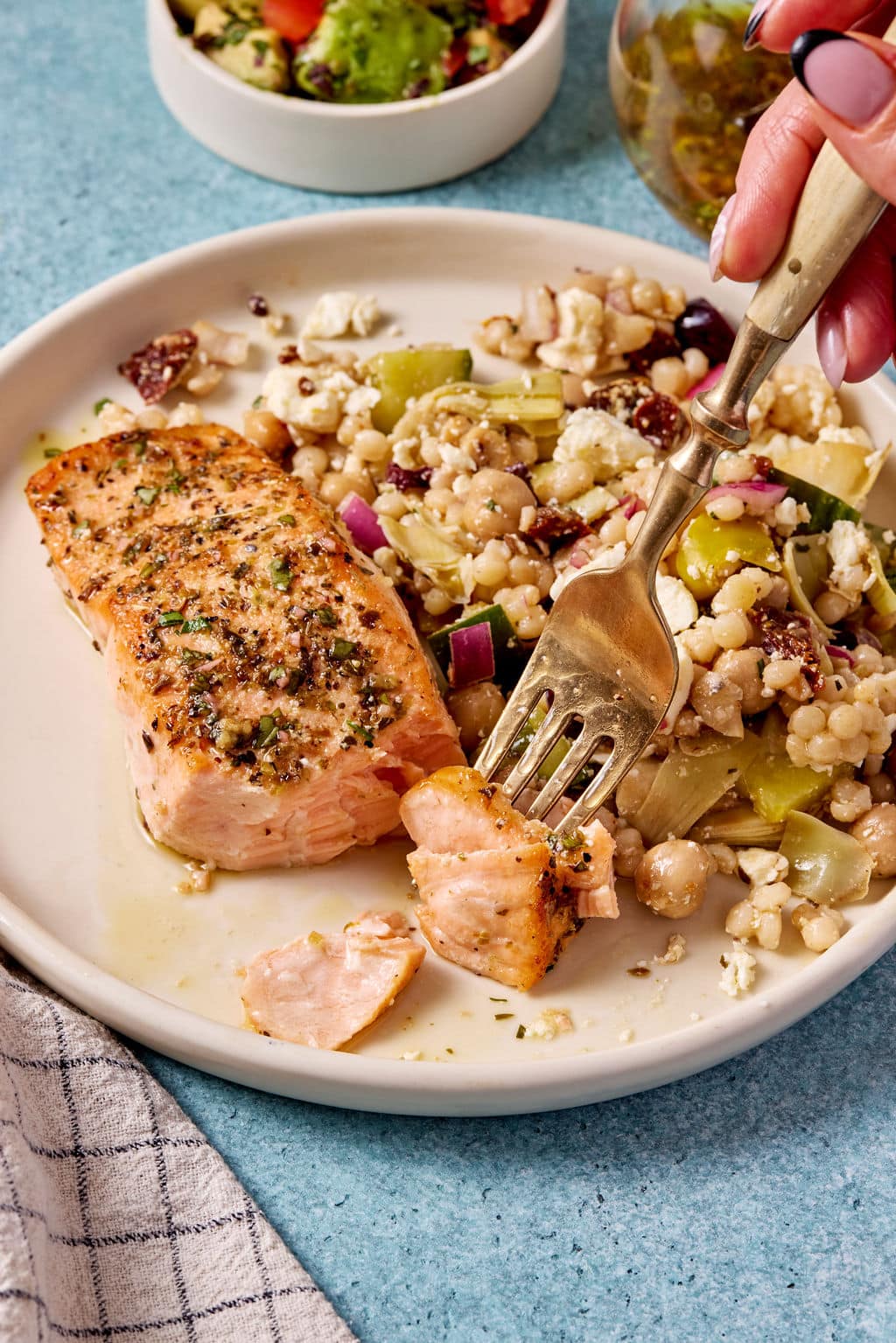 Fork lifting a flaky bite of Mediterranean salmon next to a pearl couscous salad with chickpeas, feta, cucumber, and artichokes.