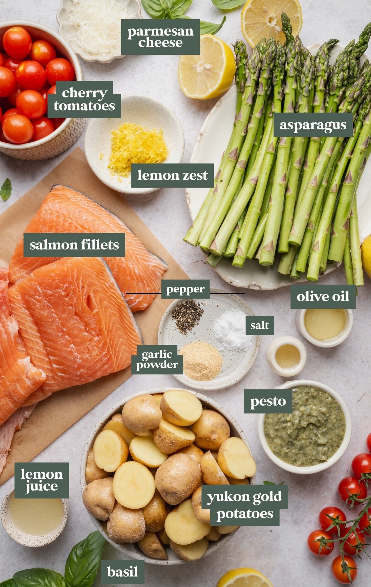 Ingredients for sheet pan pesto salmon arranged on a countertop, including salmon fillets, baby Yukon gold potatoes, asparagus, cherry tomatoes, pesto, Parmesan cheese, lemon, olive oil, garlic powder, salt, pepper, and fresh basil.