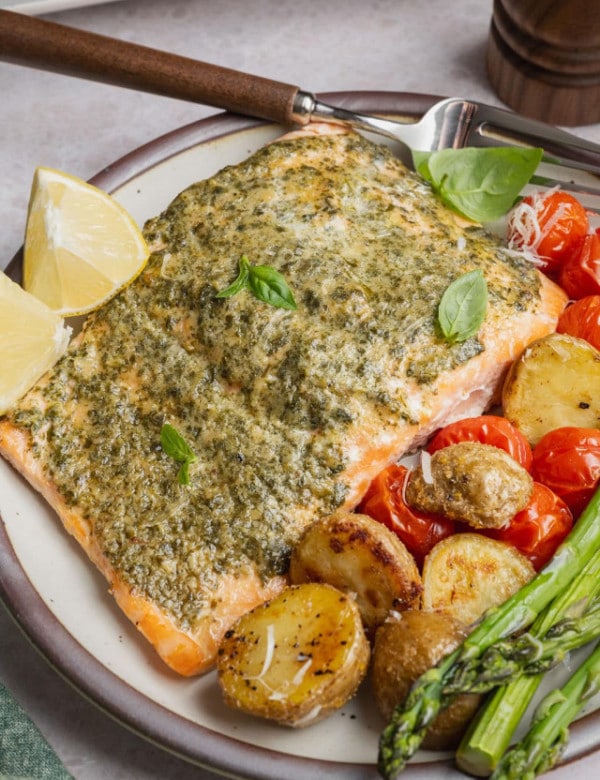 Plate of sheet pan pesto salmon served with roasted baby Yukon gold potatoes, asparagus, blistered cherry tomatoes, fresh basil, and lemon wedges.