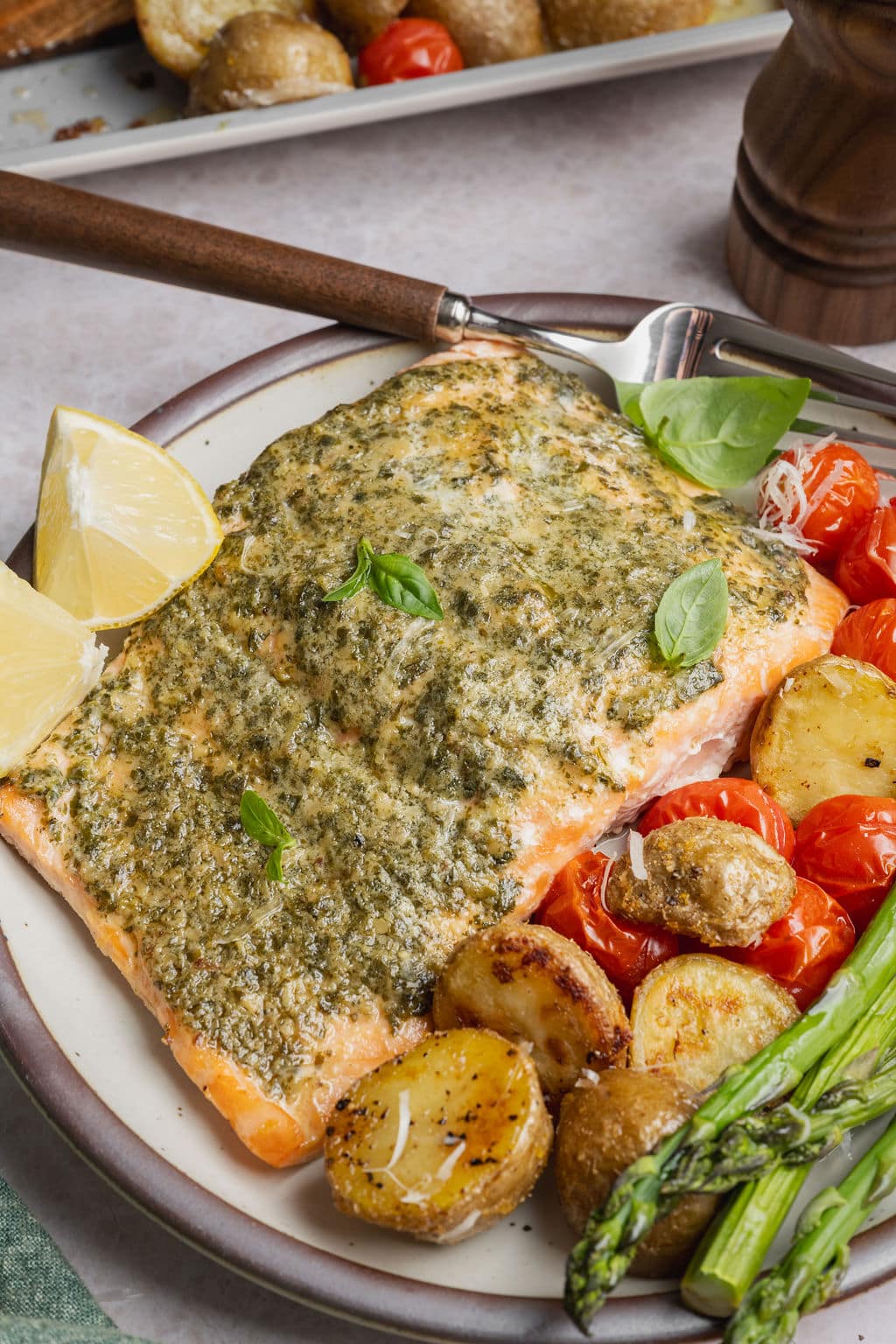 Plate of sheet pan pesto salmon served with roasted baby Yukon gold potatoes, asparagus, blistered cherry tomatoes, fresh basil, and lemon wedges.