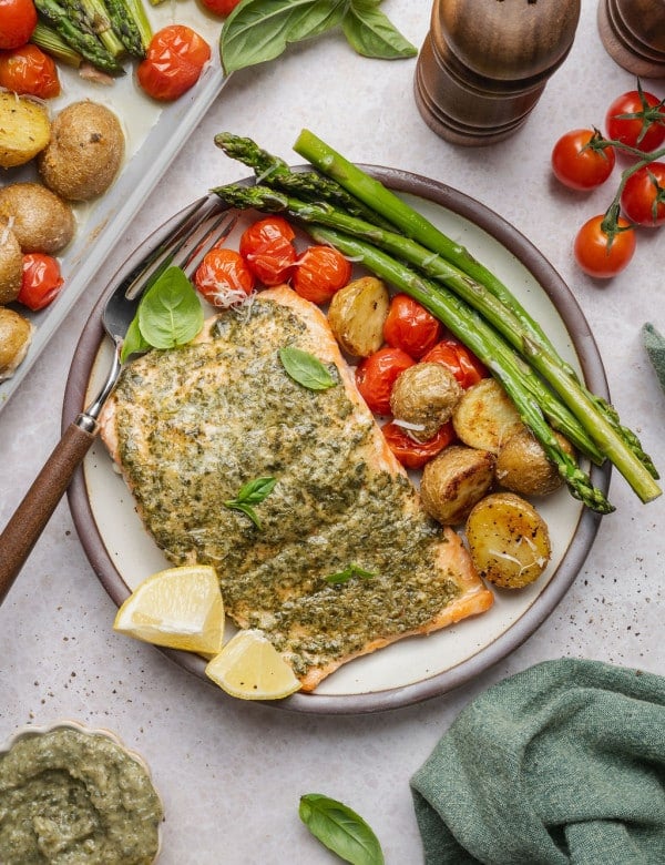 Sheet pan pesto salmon plated with roasted asparagus, crispy baby potatoes, blistered cherry tomatoes, fresh basil, grated Parmesan cheese, and lemon wedges.
