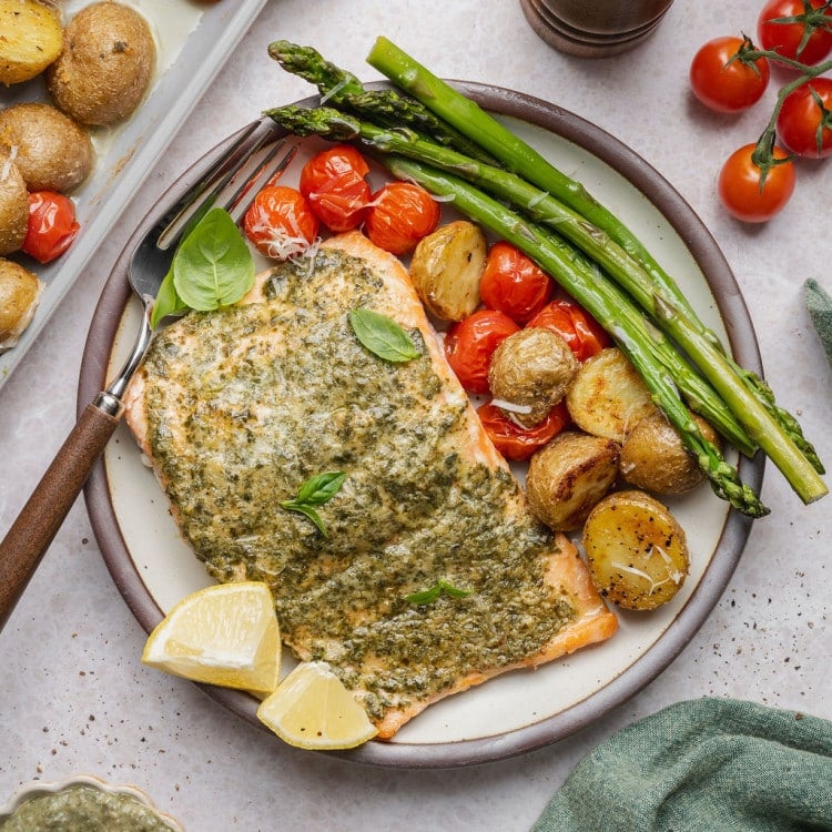 Sheet pan pesto salmon plated with roasted asparagus, crispy baby potatoes, blistered cherry tomatoes, fresh basil, grated Parmesan cheese, and lemon wedges.