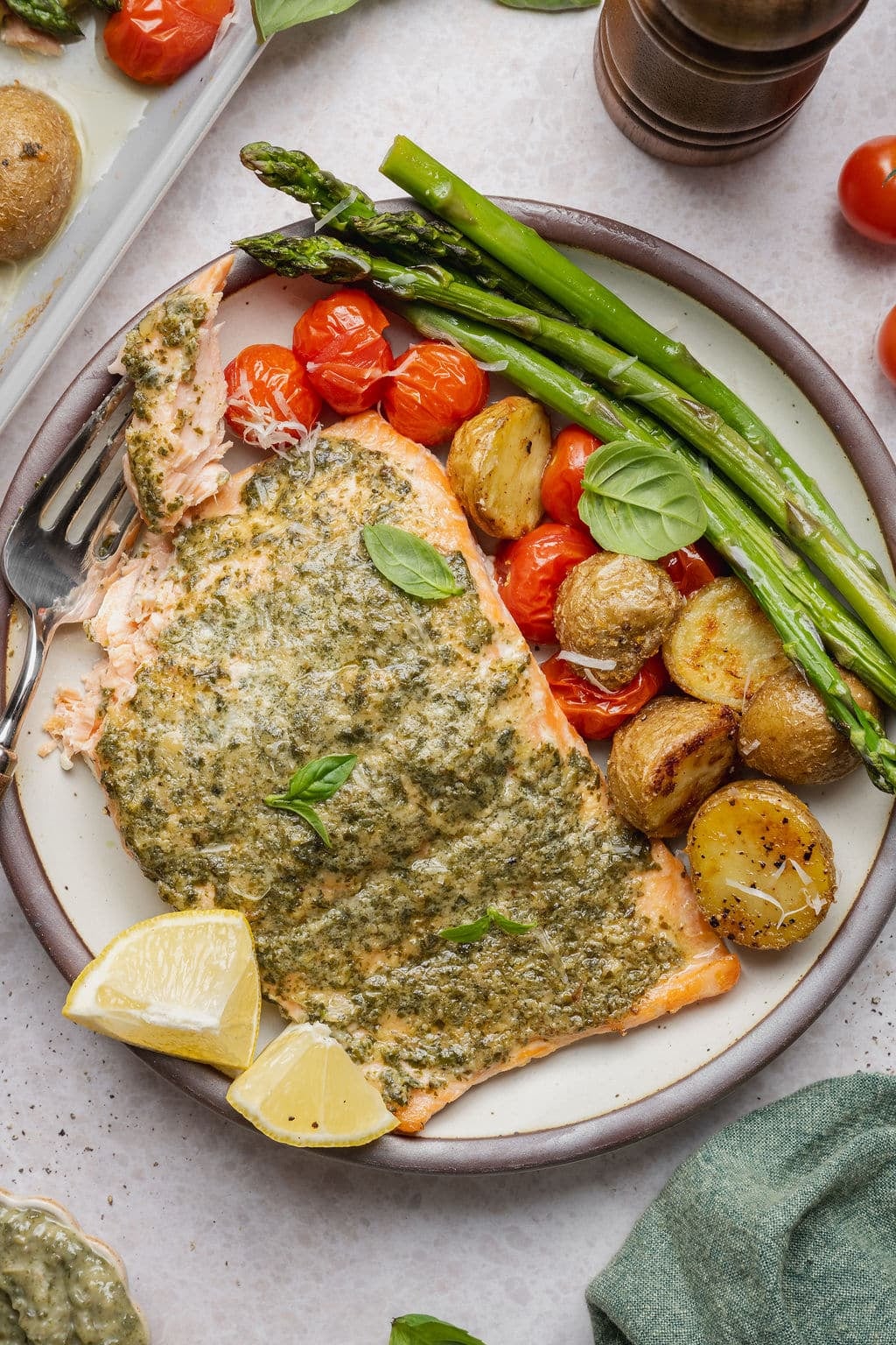 Plate of sheet pan pesto salmon with a bite removed, served with roasted asparagus, crispy baby Yukon gold potatoes, blistered cherry tomatoes, fresh basil, and lemon wedges.