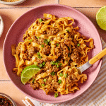 Bowl of saucy ground beef and noodles garnished with green onions, sesame seeds, chili crisp, and a lime wedge, with extra noodles in the background.