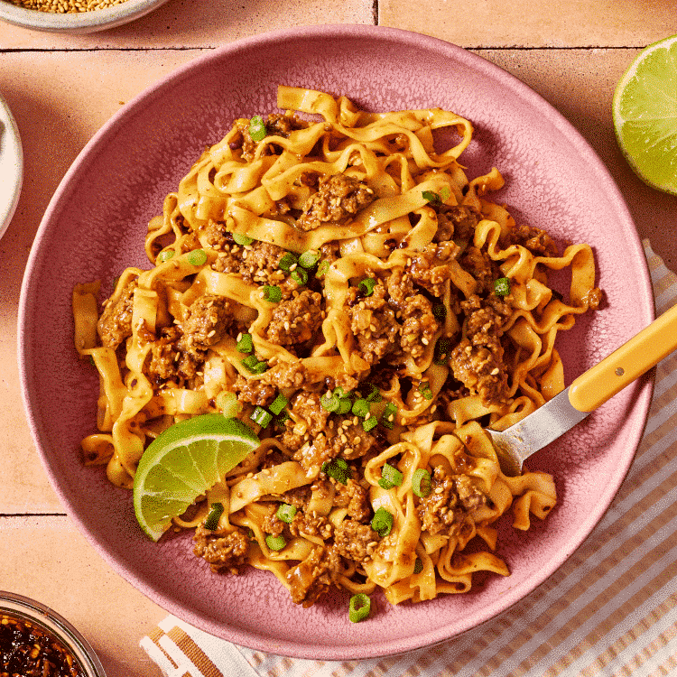 Bowl of saucy ground beef and noodles garnished with green onions, sesame seeds, chili crisp, and a lime wedge, with extra noodles in the background.