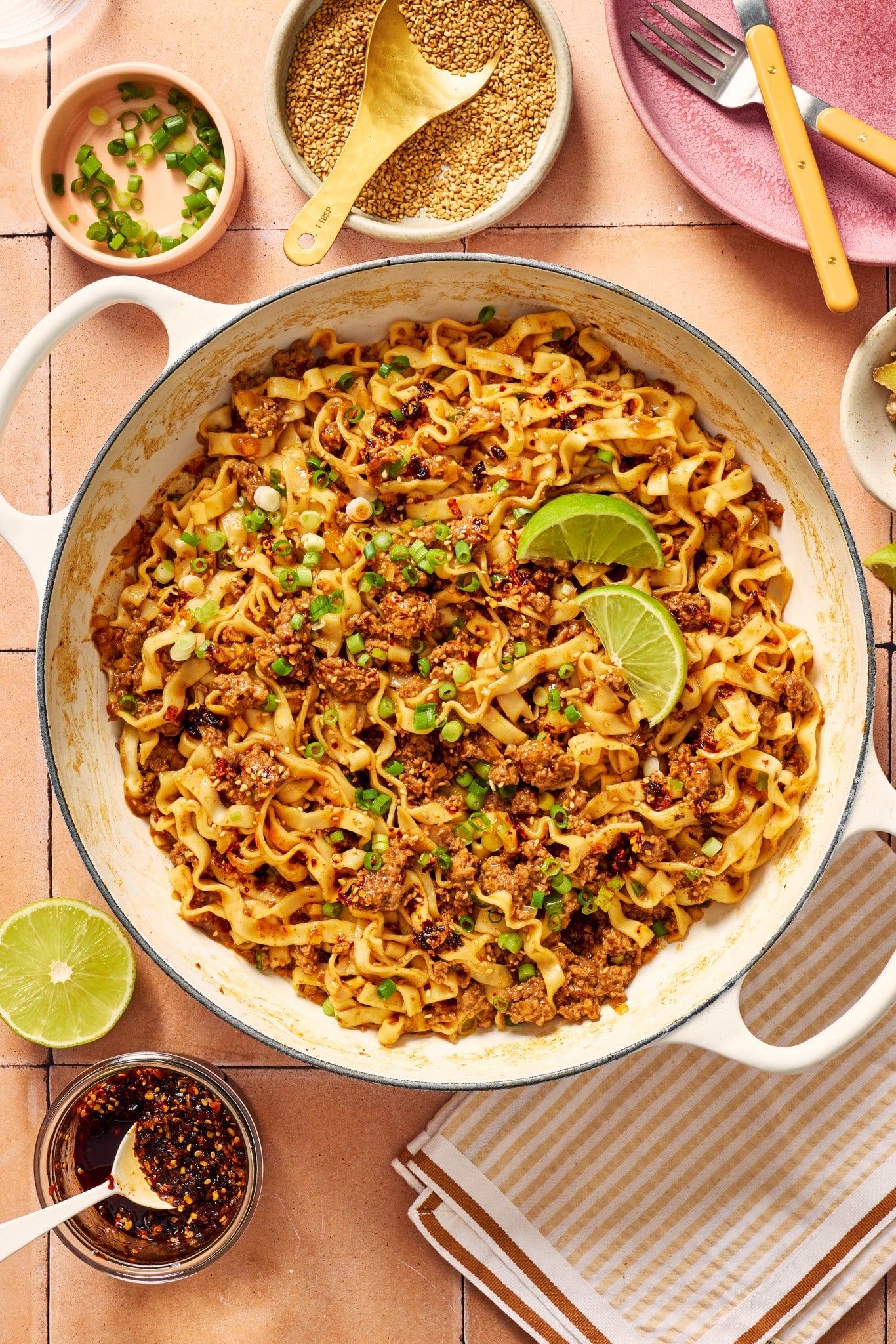 Skillet of saucy ground beef and noodles topped with green onions, sesame seeds, chili crisp, and fresh lime wedges.