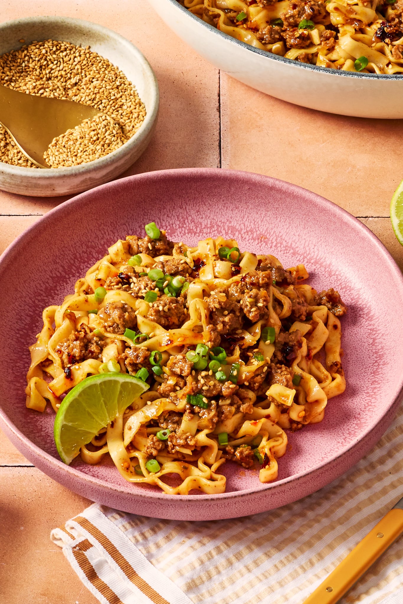 Close-up of saucy ground beef and noodles in a pink bowl with green onions, sesame seeds, chili crisp, and a lime wedge.
