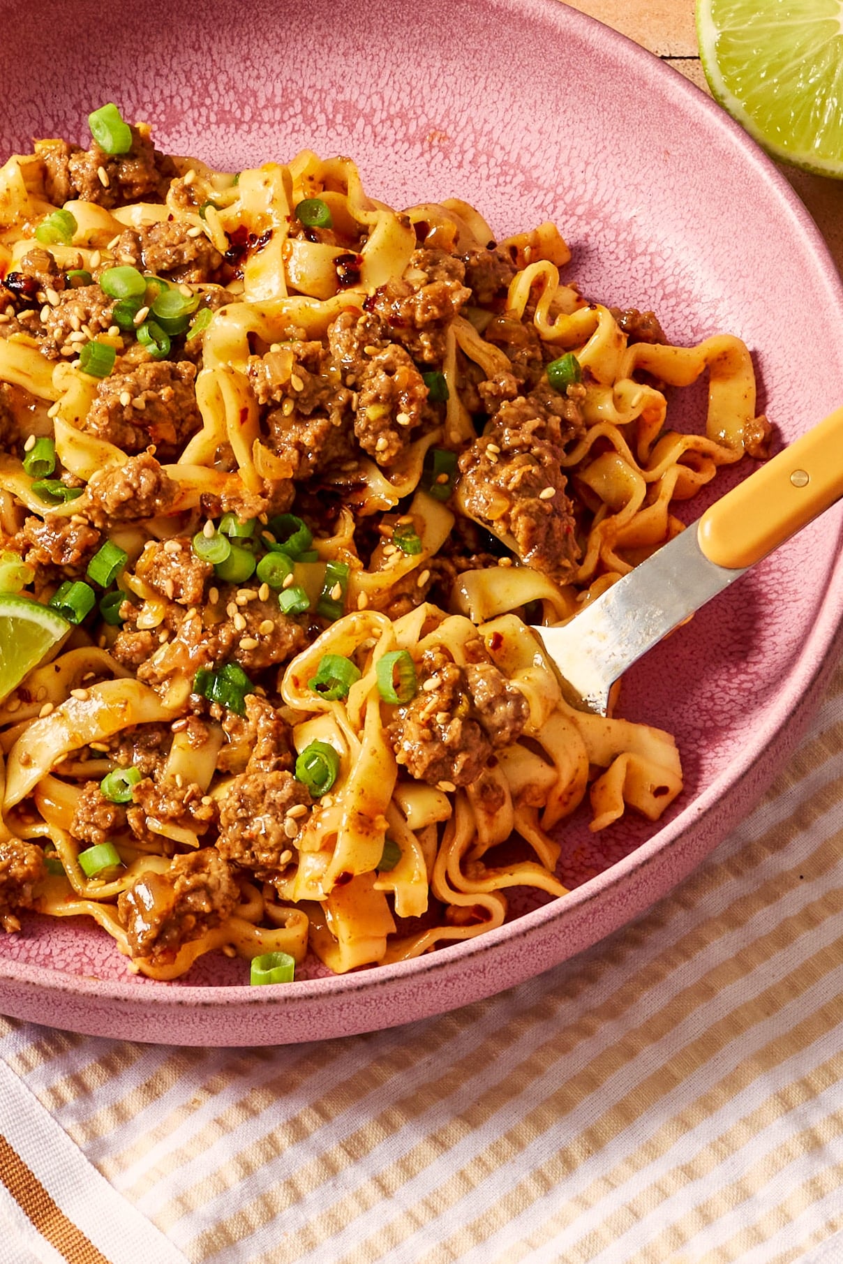 Close-up of glossy saucy ground beef and noodles topped with sliced green onions and sesame seeds, with a fork in the bowl.