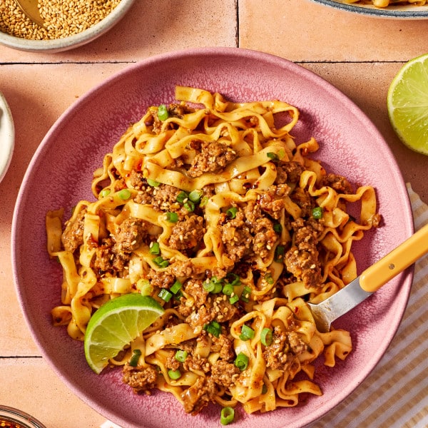 Bowl of saucy ground beef and noodles garnished with green onions, sesame seeds, chili crisp, and a lime wedge, with extra noodles in the background.