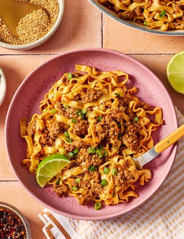 Bowl of saucy ground beef and noodles garnished with green onions, sesame seeds, chili crisp, and a lime wedge, with extra noodles in the background.