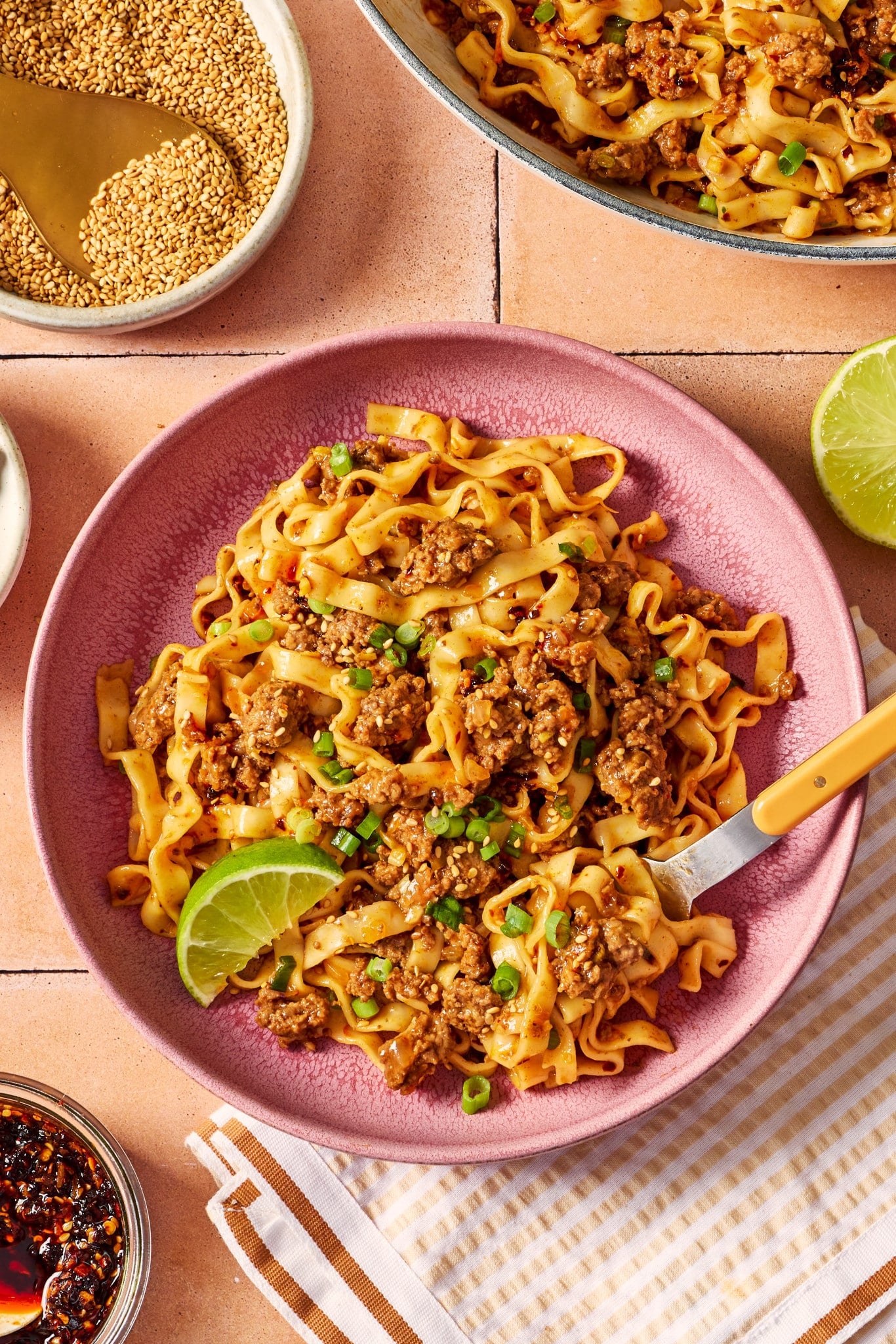 Bowl of saucy ground beef and noodles garnished with green onions, sesame seeds, chili crisp, and a lime wedge, with extra noodles in the background.