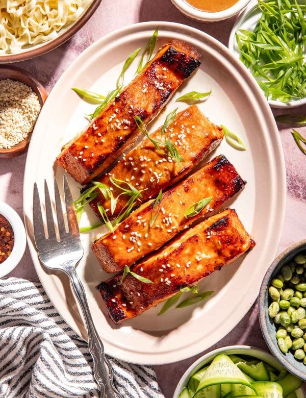 Brown sugar miso glazed salmon garnished with sesame seeds and sliced green onions, served with ramen noodles, edamame, cucumber ribbons, avocado, and extra miso glaze.