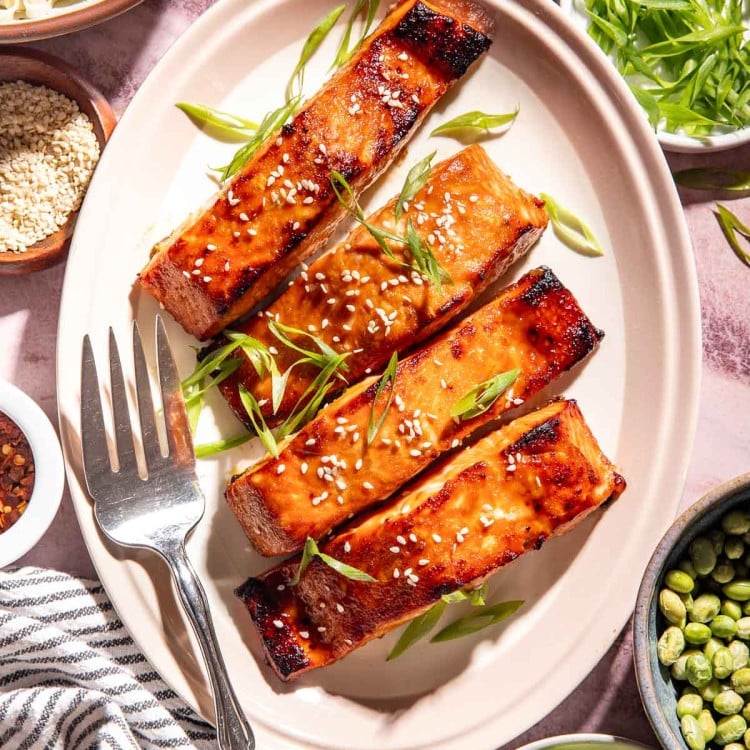 Brown sugar miso glazed salmon garnished with sesame seeds and sliced green onions, served with ramen noodles, edamame, cucumber ribbons, avocado, and extra miso glaze.