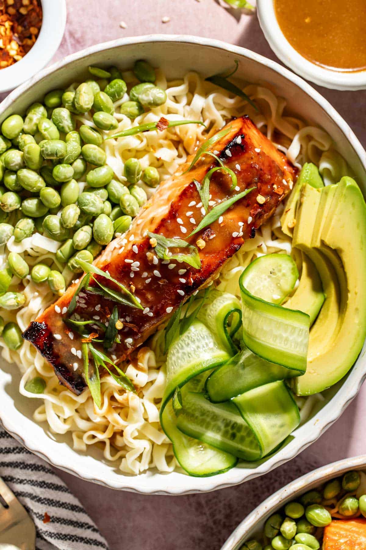 Close-up of a brown sugar miso glazed salmon noodle bowl with ramen noodles, edamame, avocado, cucumber ribbons, sesame seeds, and green onions.
