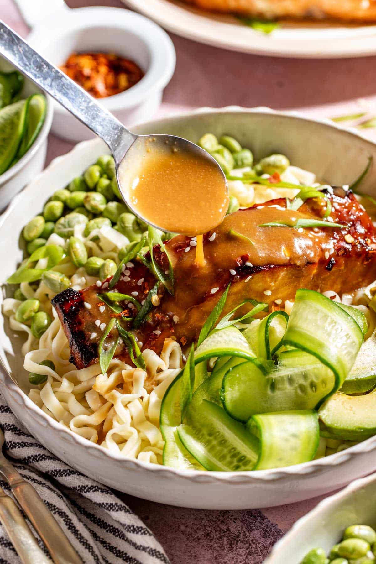 Extra brown sugar miso glaze being spooned over salmon served with ramen noodles, edamame, avocado, and cucumber ribbons.