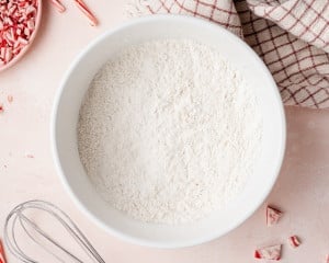 Large white mixing bowl filled with whisked dry ingredients for cookie dough, surrounded by peppermint pieces, a whisk, and a kitchen towel on a pink surface.