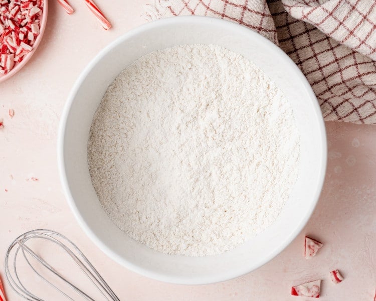 Large white mixing bowl filled with whisked dry ingredients for cookie dough, surrounded by peppermint pieces, a whisk, and a kitchen towel on a pink surface.