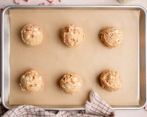 Six large scoops of peppermint cookie dough evenly spaced on a parchment-lined baking sheet, ready to bake.