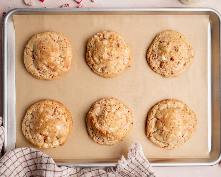 Six freshly baked white chocolate peppermint cookies cooling on a parchment-lined baking sheet.