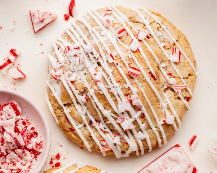 Close-up of a baked white chocolate peppermint cookie drizzled with white chocolate and topped with crushed candy cane pieces, with extra peppermint bark scattered around.