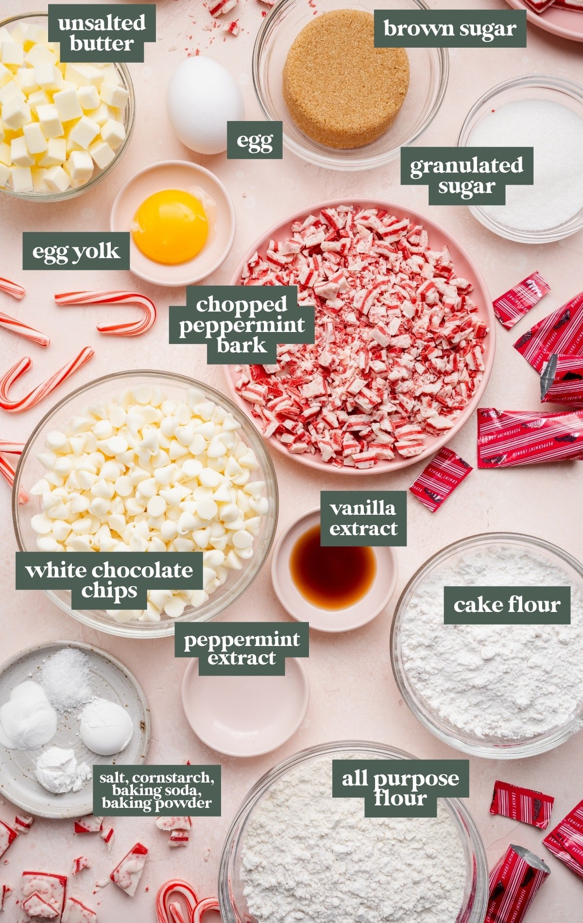 Overhead view of prepped ingredients for white chocolate peppermint cookies, including cubed butter, brown and granulated sugar, an egg and egg yolk, chopped peppermint bark, white chocolate chips, vanilla and peppermint extracts, cake flour, all-purpose flour, and small bowls of salt, cornstarch, baking soda, and baking powder.