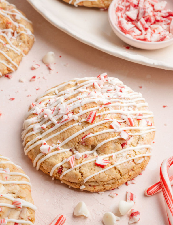 A round white chocolate peppermint cookie covered in white chocolate drizzle and crushed candy cane pieces, surrounded by scattered peppermint crumbs and candy canes.