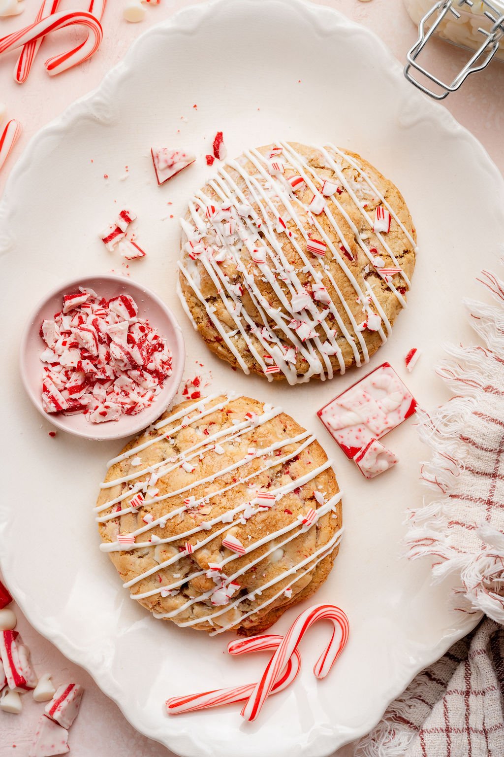 Overhead view of two large white chocolate peppermint cookies on a scalloped white platter, each drizzled with white chocolate and topped with crushed candy canes. A small pink bowl filled with chopped peppermint bark sits beside the cookies, with scattered candy cane pieces and a striped kitchen towel adding festive detail around the tray.