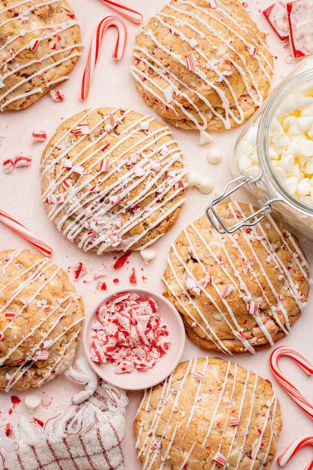 Overhead view of large white chocolate peppermint cookies drizzled with white chocolate and topped with crushed candy canes, surrounded by candy cane pieces, white chocolate chips, and a small bowl of peppermint bark on a light pink surface.