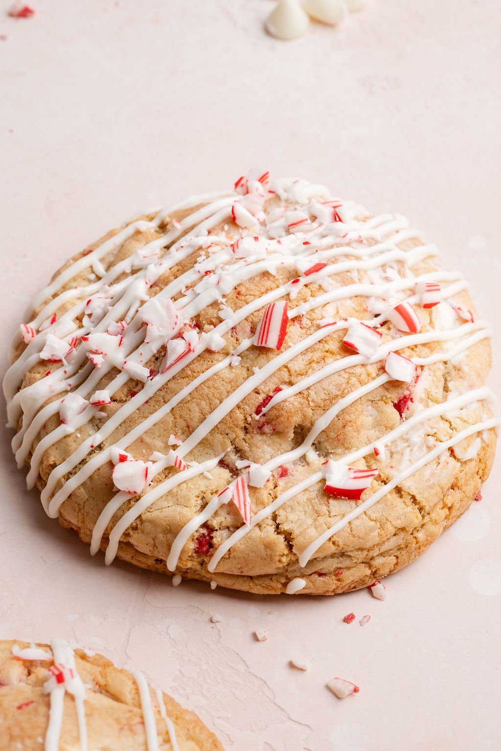 Close-up of a single white chocolate peppermint cookie topped with white chocolate drizzle and crushed candy canes on a light pink surface.