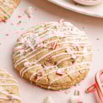 A round white chocolate peppermint cookie covered in white chocolate drizzle and crushed candy cane pieces, surrounded by scattered peppermint crumbs and candy canes.