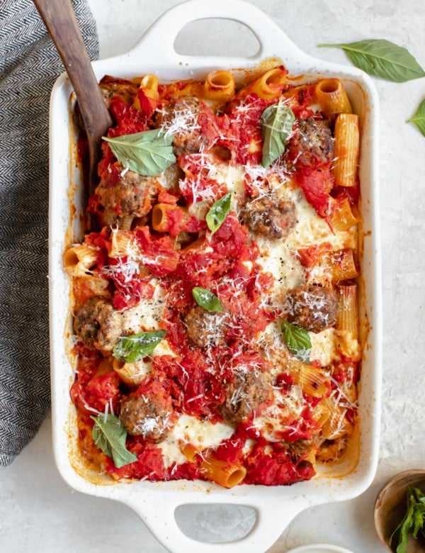 baked meatballs with rigatoni pasta in a white casserole dish topped with basil and parmesan cheese