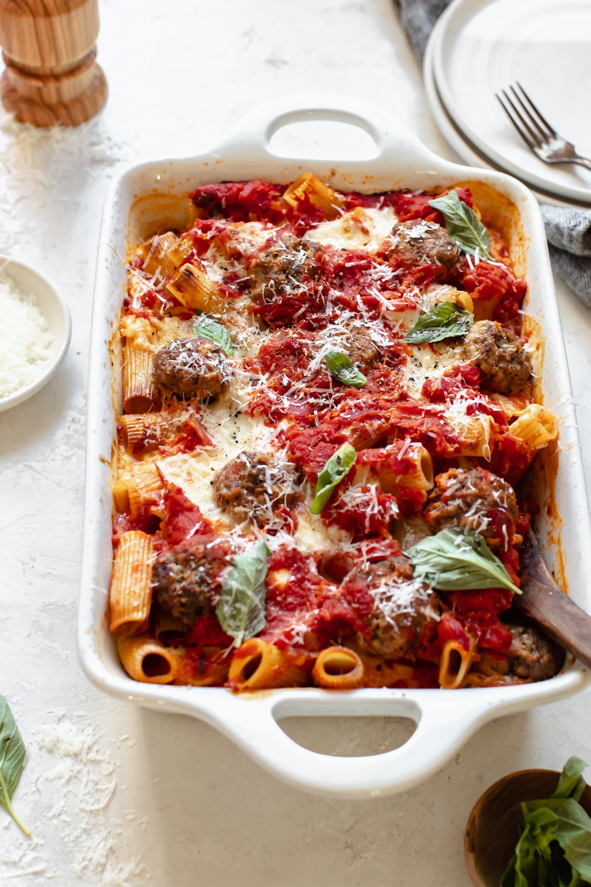 baked meatballs with rigatoni pasta in a white casserole dish topped with basil and parmesan cheese