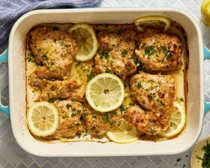 Overhead view of baked Greek yogurt lemon chicken in a casserole dish, garnished with lemon slices, parsley, and lemon zest.