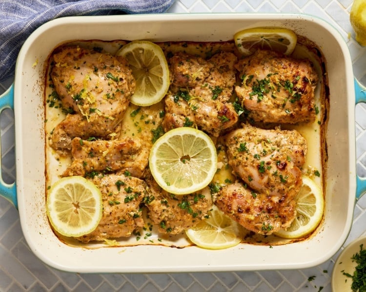 Overhead view of baked Greek yogurt lemon chicken in a casserole dish, garnished with lemon slices, parsley, and lemon zest.