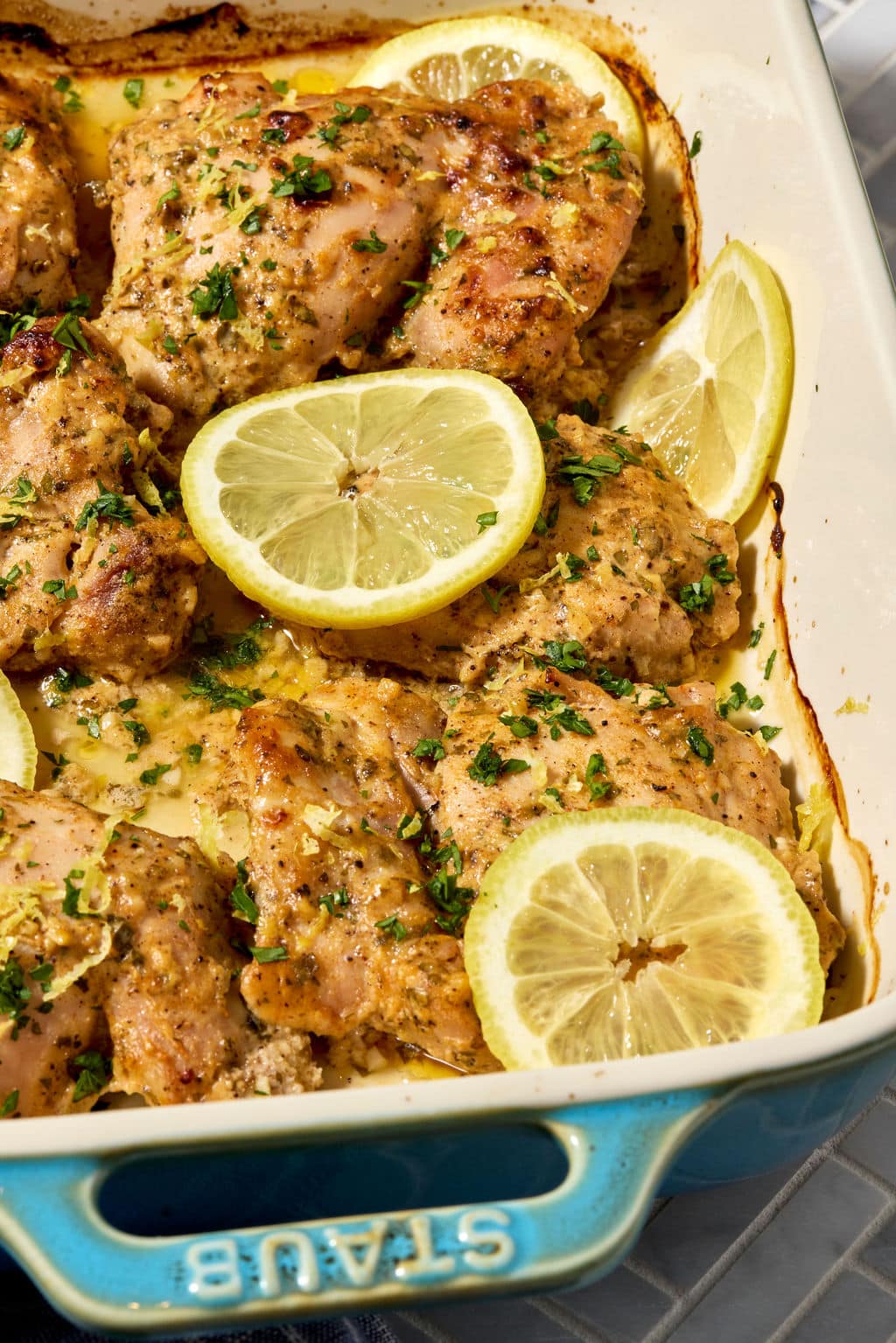 Close-up of baked Greek yogurt lemon chicken thighs in a white baking dish, garnished with fresh parsley, lemon zest, and lemon slices, with golden pan juices surrounding the chicken.