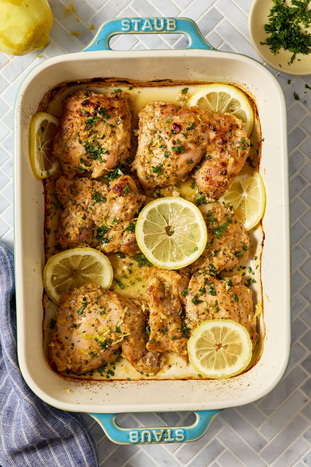 Overhead view of Greek yogurt lemon chicken baked in a white casserole dish with lemon slices, fresh parsley, and lemon zest, showing lightly browned chicken thighs in a creamy lemony sauce.
