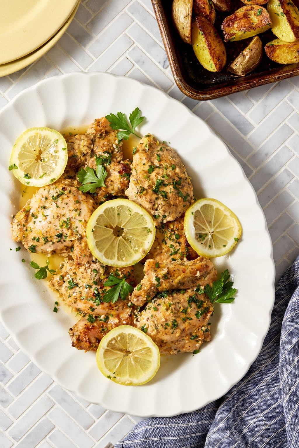 Greek yogurt lemon chicken arranged on a white serving platter and garnished with lemon slices, parsley, and lemon zest, with roasted potatoes visible in the background.