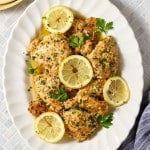 Platter of baked Greek yogurt lemon chicken topped with fresh parsley, lemon zest, and lemon slices, served with flavorful pan juices spooned over the chicken.