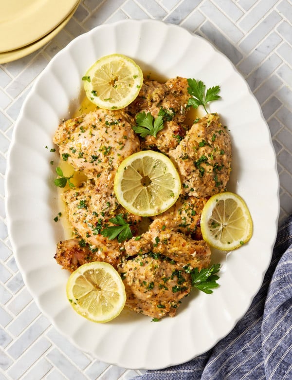 Platter of baked Greek yogurt lemon chicken topped with fresh parsley, lemon zest, and lemon slices, served with flavorful pan juices spooned over the chicken.